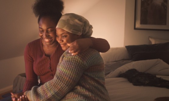 Two women (we can suppose that one is suffering from cancer) hugging, seated on a bed. 